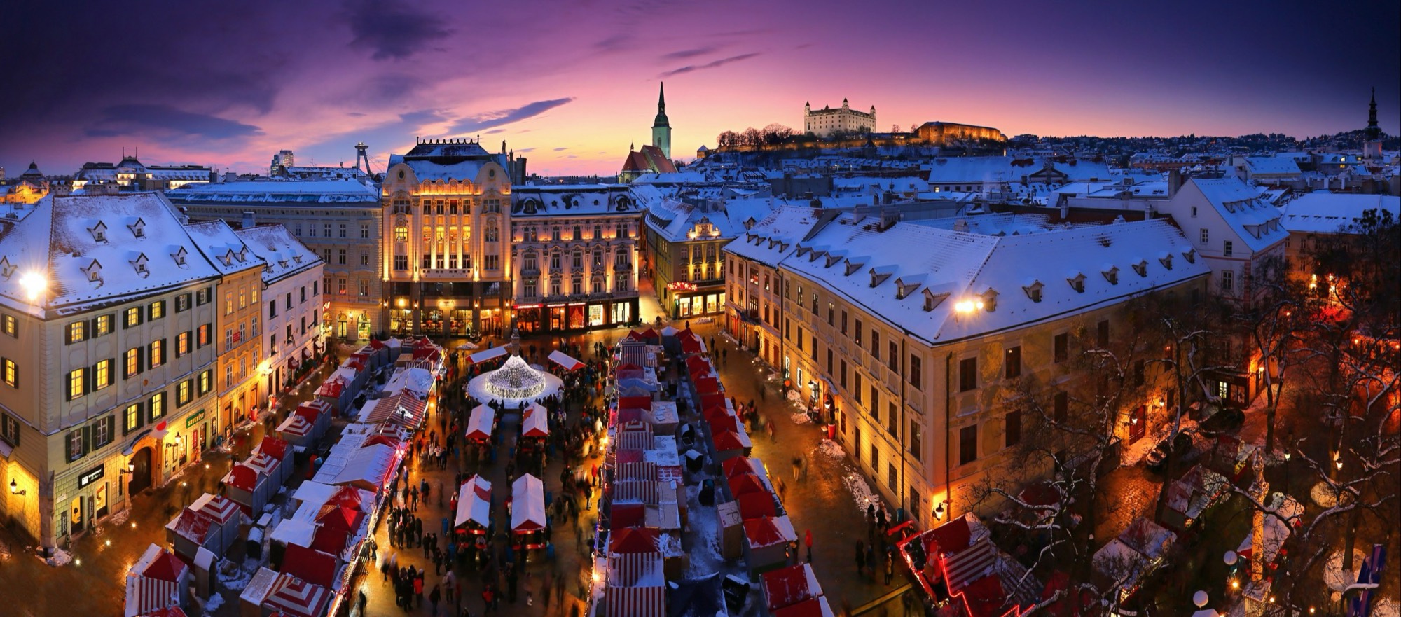 Il mercatino di Natale in Hlavné námestie al crepuscolo, visto dall’alto: bancarelle bianche e rosse nella neve, un albero illuminato e il castello sulla collina alle spalle
